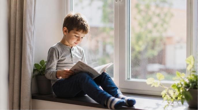 Мальчик со спокойным характером читает книгу, сидя на подоконнике